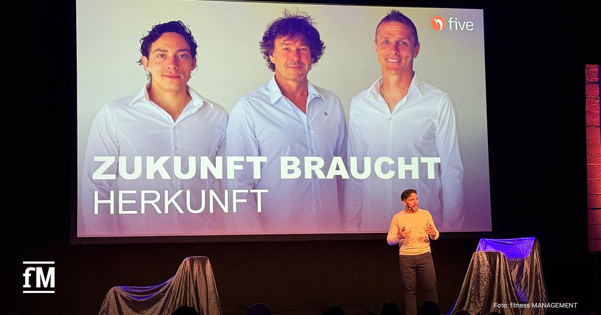 Bühne mit Vortrag, vorn ein Sprecher, dahinter große Leinwand mit drei Personen, five Logo und Schriftzug 'Zukunft braucht Herkunft', Publikum im Vordergrund.