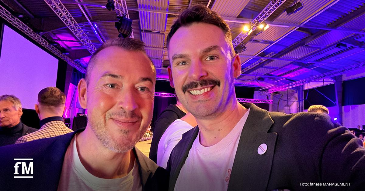 Zwei Personen (Peter Hinojal und Janosch Marx) machen ein Selfie in einer Messehalle, im Hintergrund Bühne, Lichttraversen und weitere Gäste.