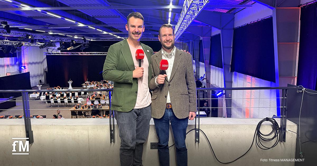 Zwei Personen (Janosch Marx & Wolfgang Packi) stehen auf einer Empore mit roten Mikrofonen, hinter ihnen eine große Halle mit gedeckten Tischen.