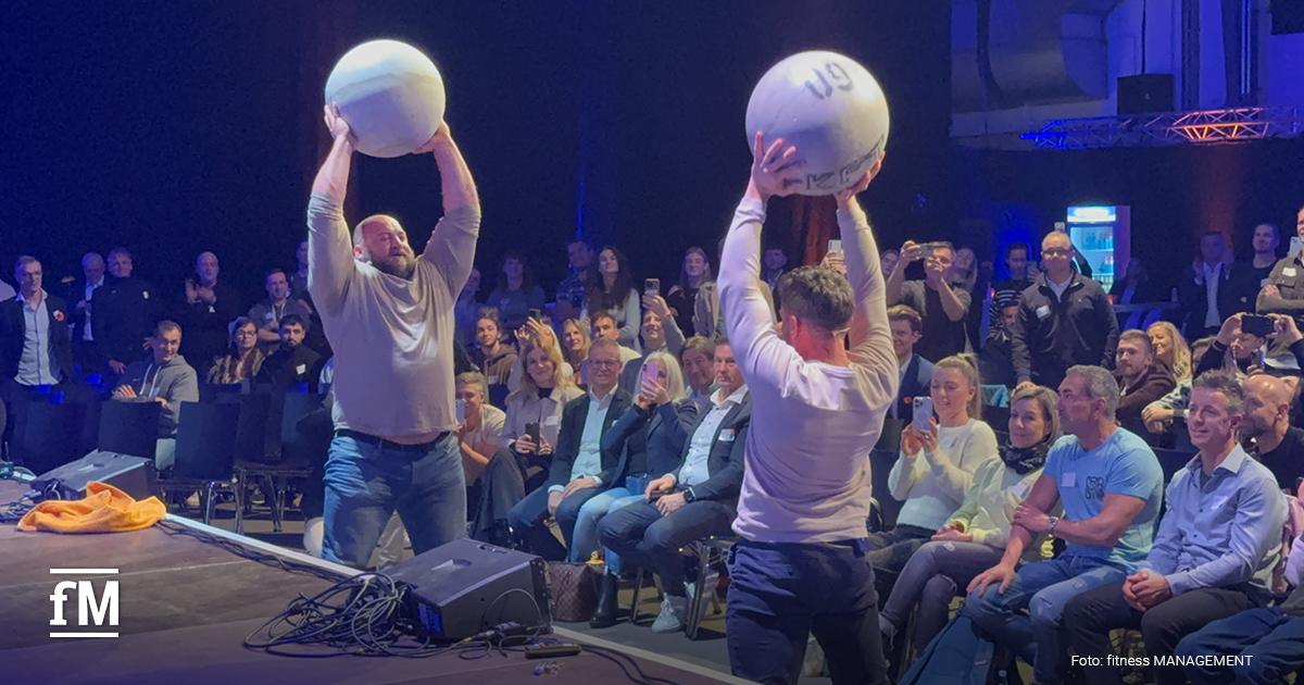 Zwei Personen (Wolf Harwarth & Dennis Kohlruss) heben große Kugeln über den Kopf auf einer Bühne, vor ihnen sitzt ein dichtes Publikum.
