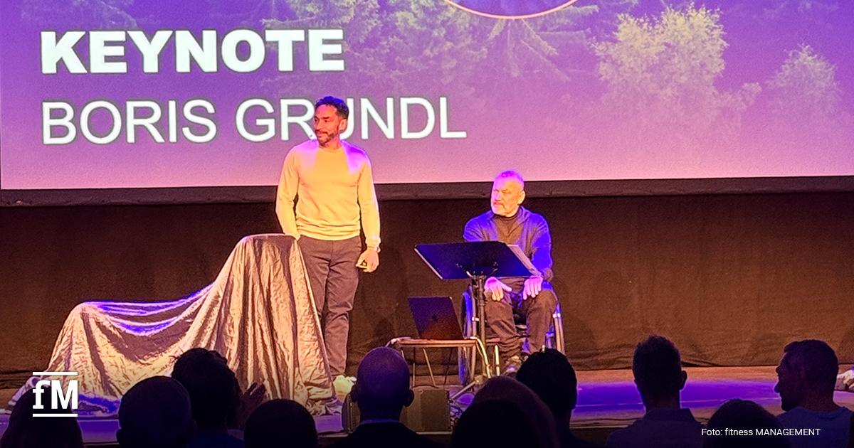 Eine Person (Wolf Harwath) steht, eine Person (Boris Grundl) sitzt im Rollstuhl auf der Bühne, dahinter Leinwand mit großem Schriftzug 'Keynote'.