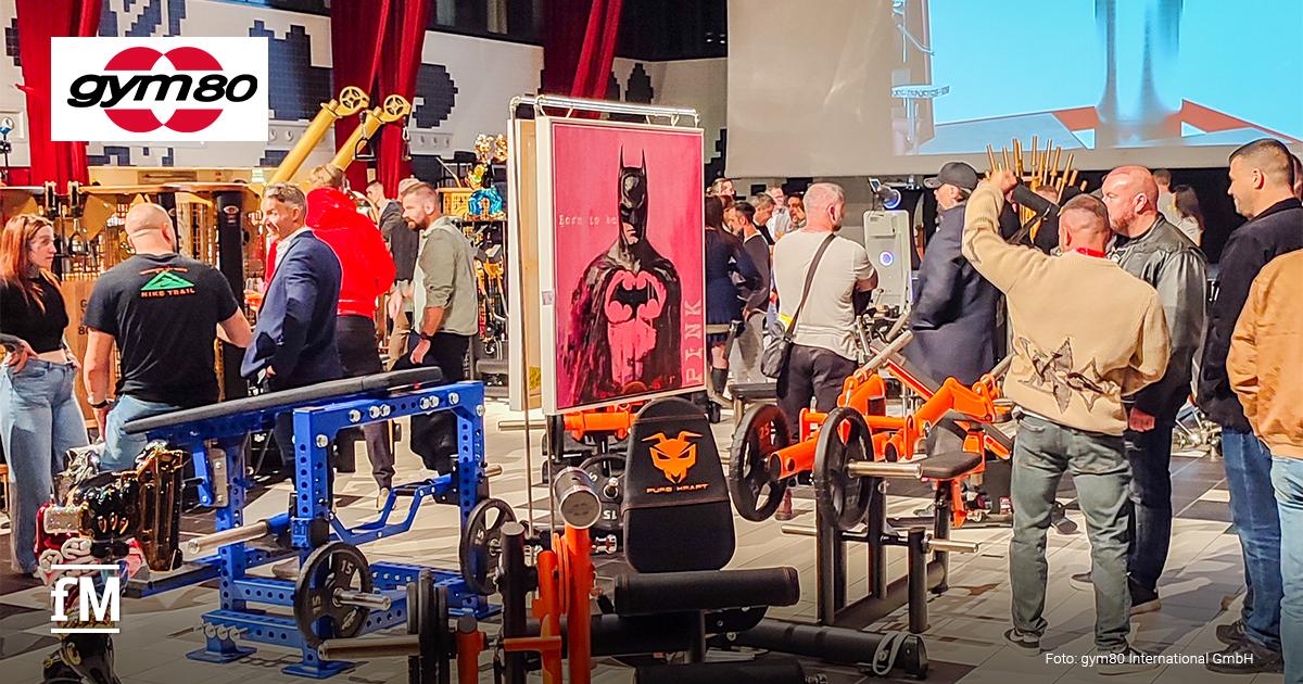 Kraft trifft Kunst 5 von gym80 Menschen betrachten Fitnessgeräte und Kunstwerke in einer großen Halle mit roten Vorhängen.