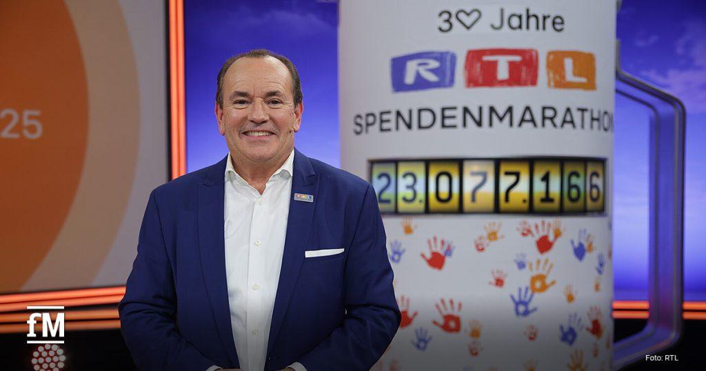 Ein Mann im blauen Anzug steht im Studio vor einer großen RTL-Spendenmarathon-Säule mit digital angezeigter Spendensumme und bunten Handabdrücken.