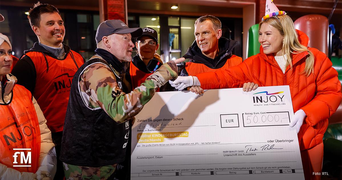 INJOY-Team mit Joey Kelly übergibt Spendencheck Mehrere Personen mit INJOY-Westen stehen um Markenbotschafter Joey Kelly und halten einen großen Scheck mit der Spendensumme von 50.000 Euro in die Kamera.
