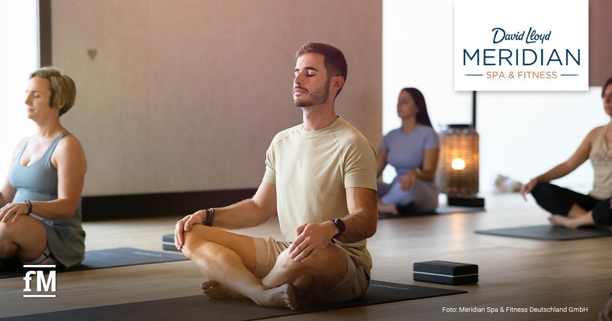 Mentale Männergesundheit DLM Weltmännertag In einem hellen Kursraum sitzen mehrere Personen im Schneidersitz auf Yogamatten und haben die Arme locker auf den Oberschenkeln liegen. Rechts oben ist das Logo von David Lloyd Meridian.