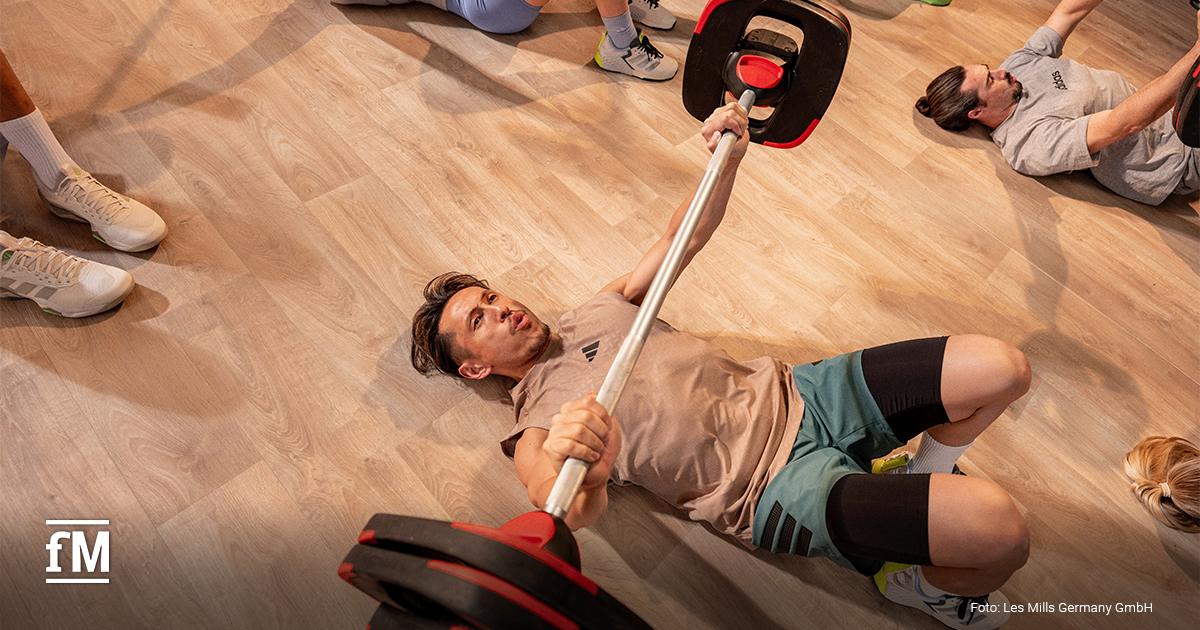 Person liegt am Boden und hebt eine Langhantel mit roten Gewichtsscheiben in einem Fitnesskurs.