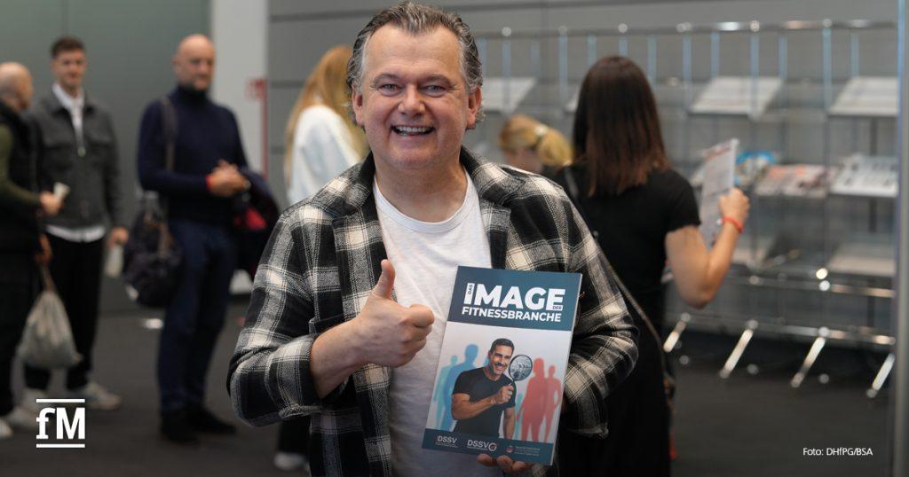 Mann im Karohemd steht im Foyer, hält lächelnd eine Studie mit der Aufschrift „Das Image der Fitnessbranche“ in die Kamera.