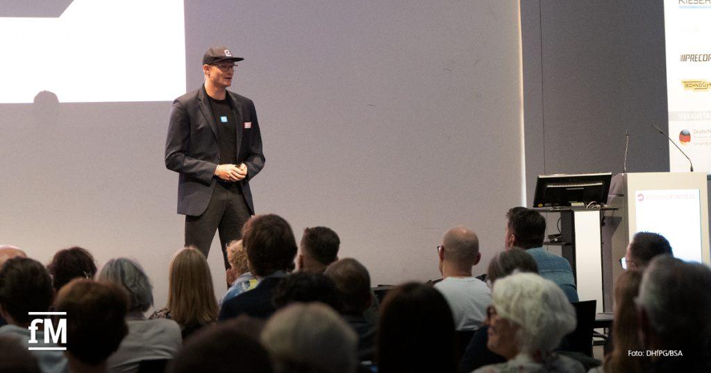 Mann mit Kappe und Sakko steht auf einer Bühne vor sitzendem Publikum in einem Vortragssaal.