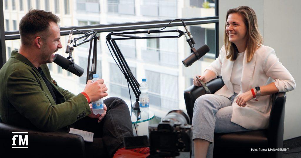 Zwei Personen sitzen sich in Sesseln gegenüber, sprechen in Mikrofone, vor einem großen Fenster mit Stadtblick.