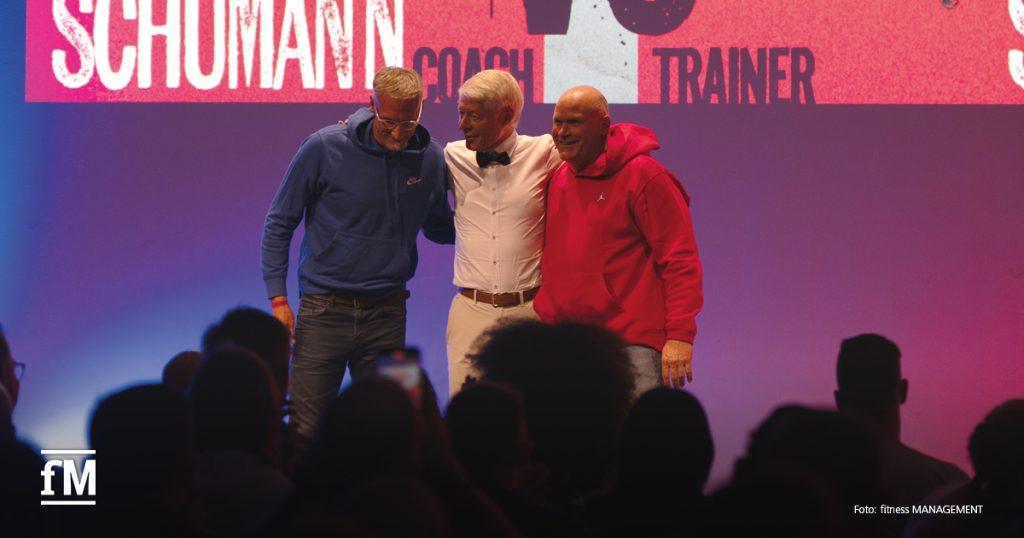 Drei Männer stehen nebeneinander auf einer beleuchteten Bühne, Publikum im Vordergrund, bunte Schrift im Hintergrund.