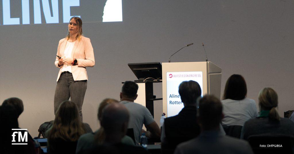 Frau im Blazer steht mit Mikrofon auf einer Bühne, rechts ein Rednerpult mit Aufstiegskongress-Logo, Publikum im Vordergrund.