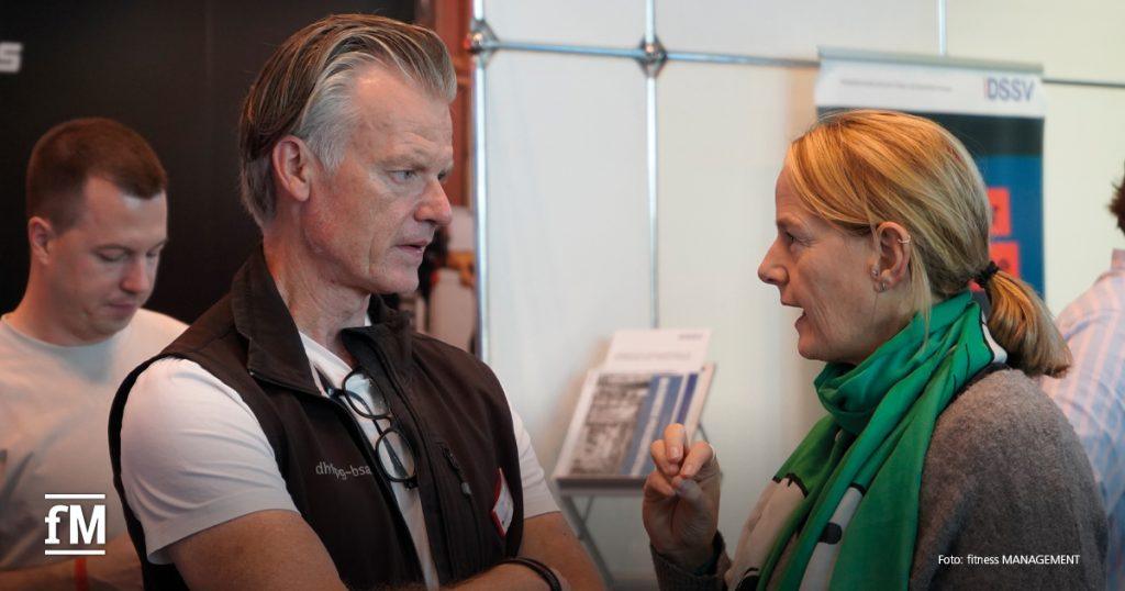 Zwei Personen stehen sich an einem Messestand gegenüber und sprechen intensiv, im Hintergrund weitere Teilnehmende.