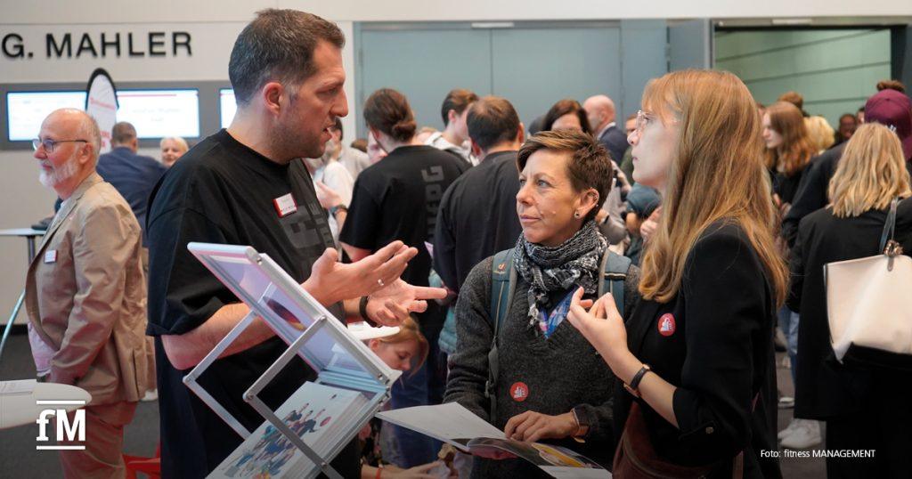 Ein Hochschulmitarbeiter berät zwei Besucherinnen an einem Stand, im Hintergrund viele Menschen.