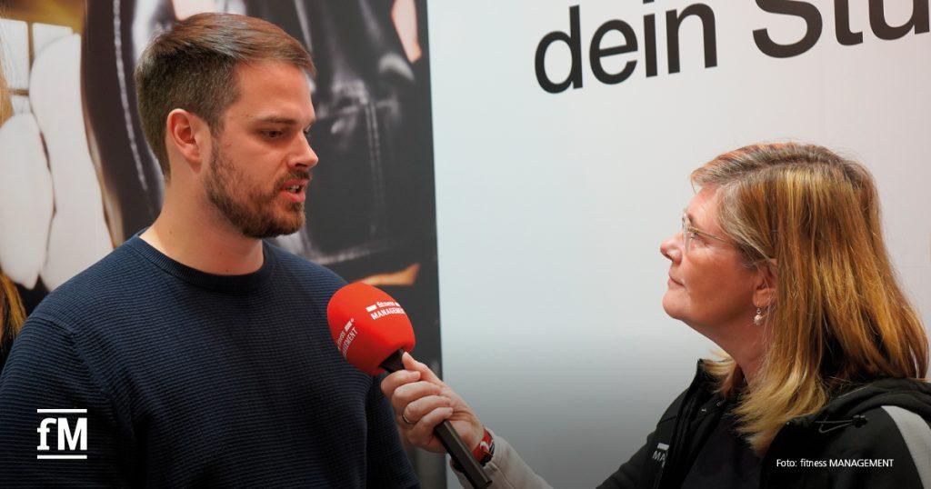 Eine Redakteurin hält ein rotes Mikrofon mit fitness-MANAGEMENT-Logo zu einem Gesprächspartner vor Interviewwand.