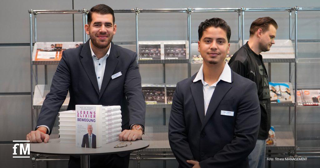 Zwei junge Männer im Anzug stehen hinter einem runden Tisch mit gestapelten Büchern, im Hintergrund Regale mit Fachmaterial.