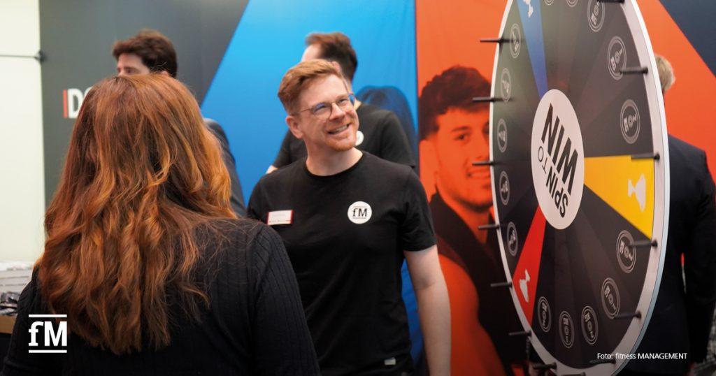 Ein Besucher steht vor einem bunten Glücksrad, ein fM-Mitarbeiter mit schwarzem Shirt lächelt ihn an, im Hintergrund Messestand.