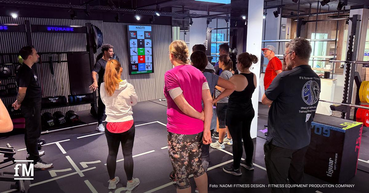 Eine Gruppe von Erwachsenen steht auf einer Trainingsfläche und hört einem Coach vor einem Bildschirm mit Workout-Grafiken zu.