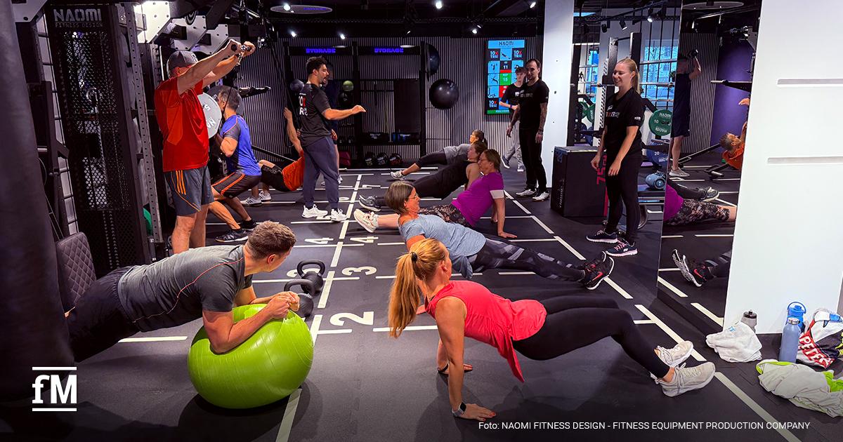 Auf der markierten Trainingsfläche trainieren mehrere Personen gleichzeitig mit Bällen, Kettlebells und Rigs, während Coaches sie anleiten.