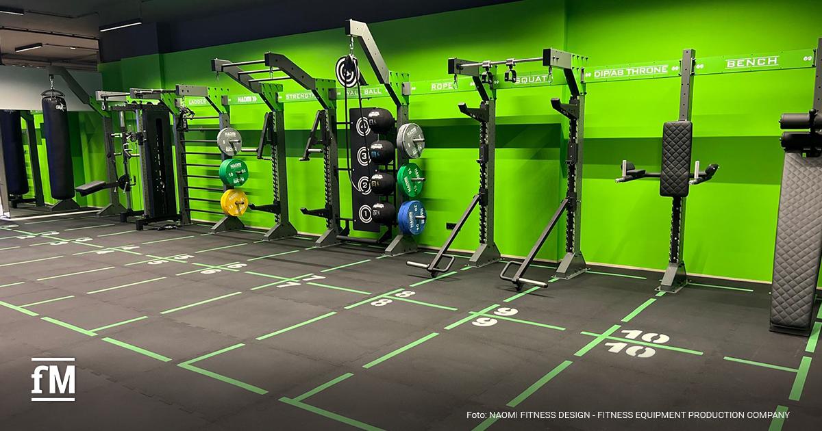 Breite grüne Wand mit mehreren Trainingsracks, Boxsack und einem mit Zahlen von eins bis zehn markiertem schwarzen Boden in einem Studio.