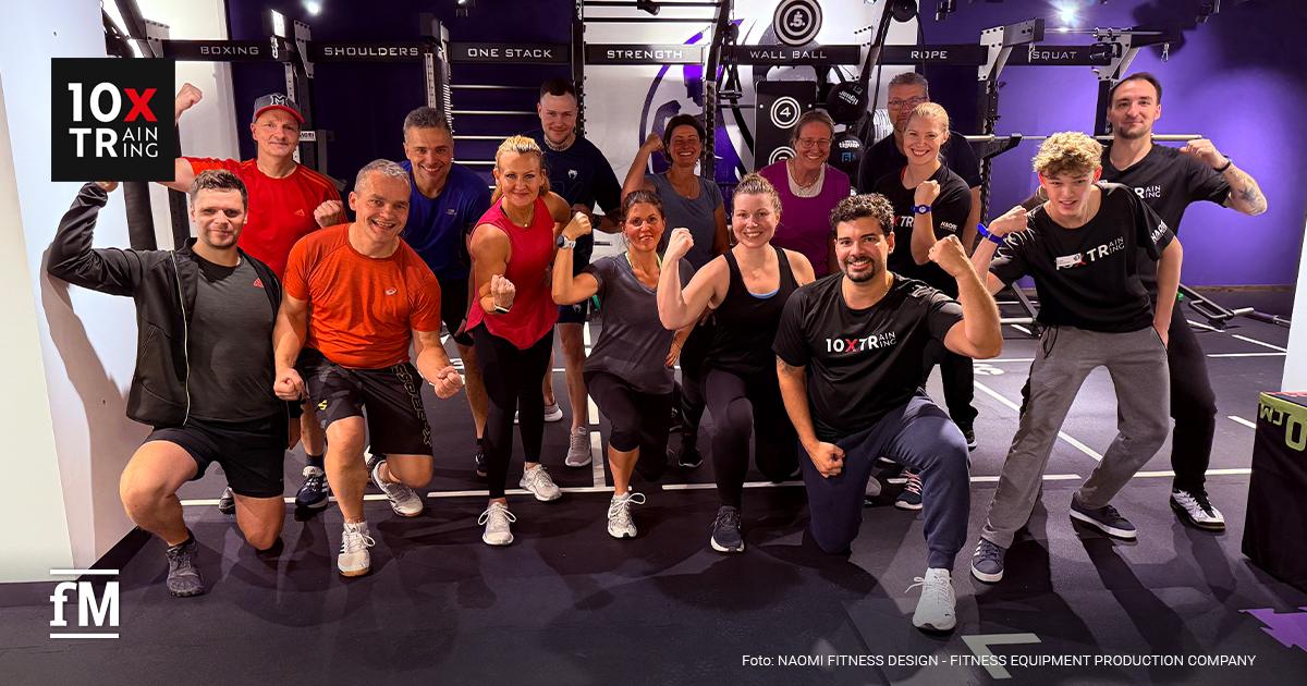 Naomi Fitness Design 10XTraining Gruppe von etwa fünfzehn Personen kniet und steht in einem Fitnessstudio und posiert lachend vor einem Trainingsrack.