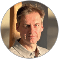 Porträt eines Mannes mit kurzem grauem Haar in einem hellgrauen Polohemd, der in warmem Licht in die Kamera blickt. Er steht vor einem neutralen Hintergrund.