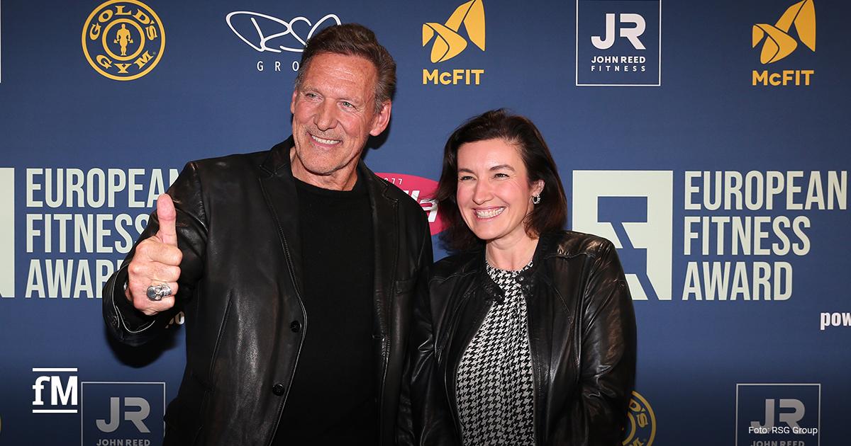 Zwei Personen (Dorothee Bär und Ralf Moeller) in dunkler Kleidung stehen lächelnd vor einer blauen Fotowand mit Logos des European Fitness Award und Partnern.