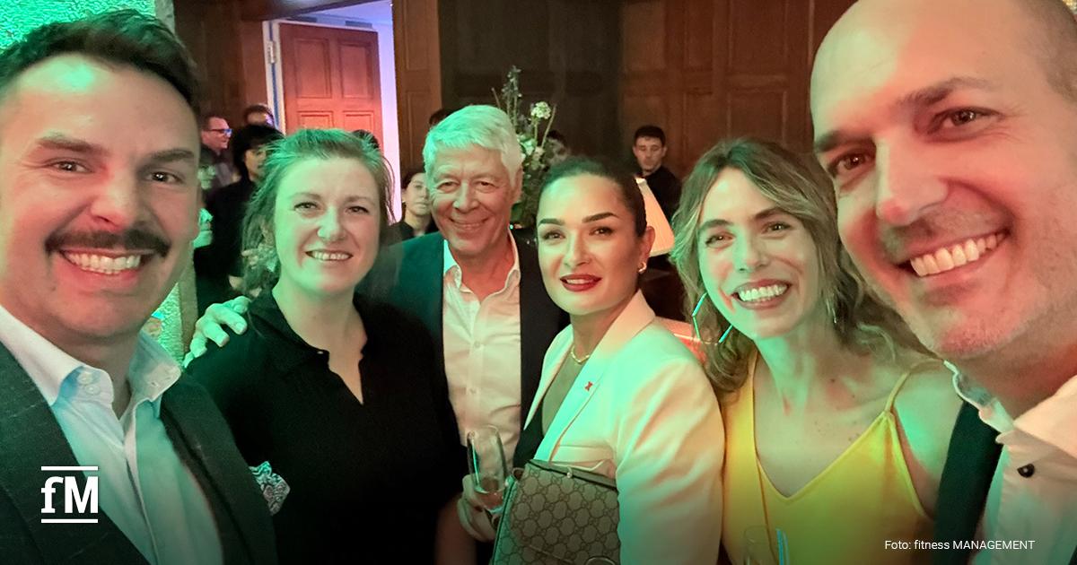 Selfie von sechs Personen in Abendkleidung, die dicht nebeneinander in einem festlich beleuchteten Raum in die Kamera lächeln.