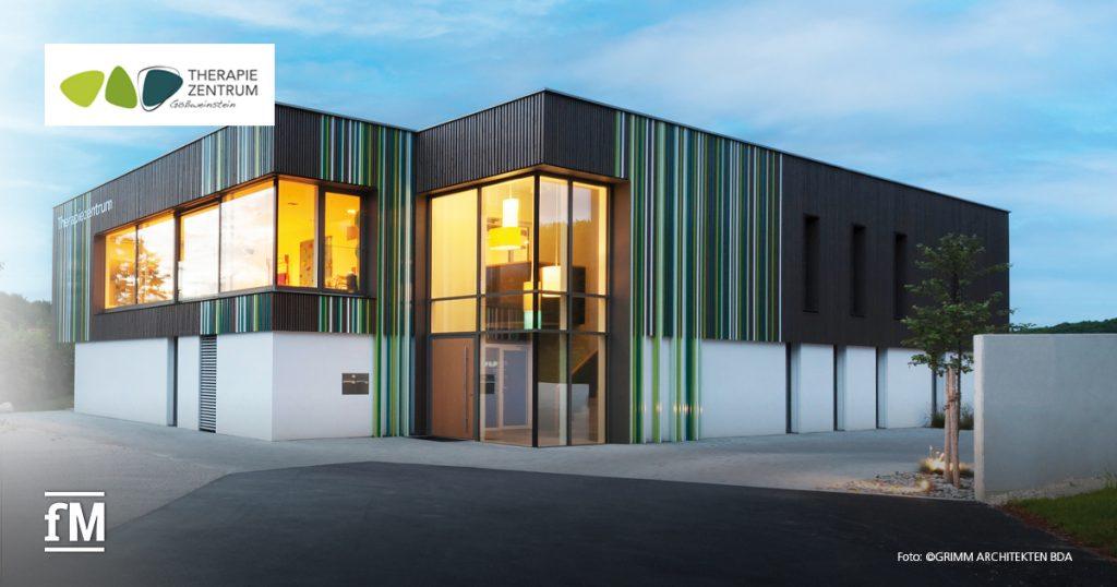 Moderne Gebäudeansicht mit großen Fenstern und bunter Fassade bei Abendlicht, Logo des Therapiezentrums sichtbar.