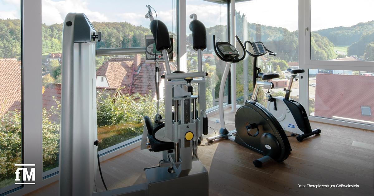 Zwei Fahrradergometer und ein Kraftgerät stehen vor großen Fenstern mit Aussicht auf Dächer und bewaldete Hügel.