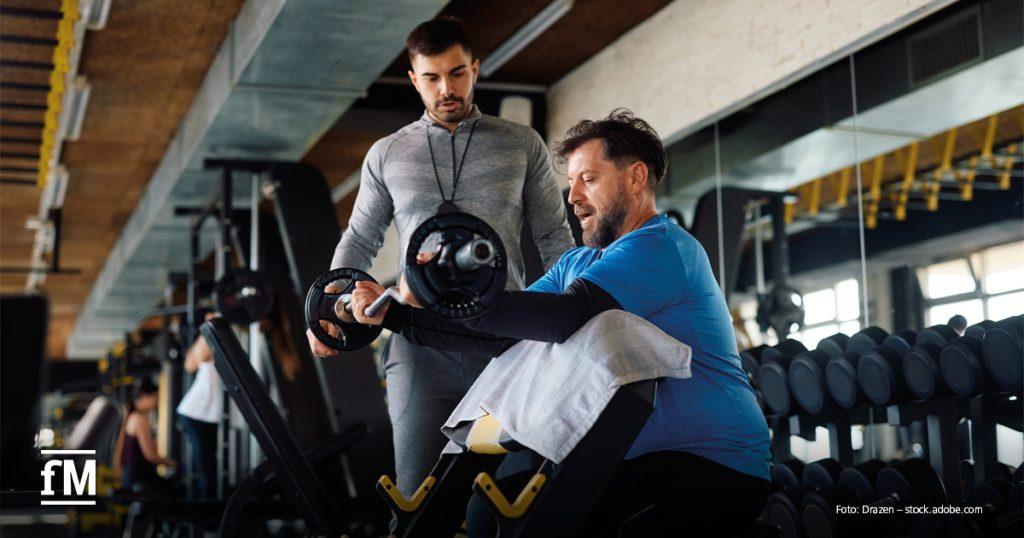 Personal Trainer unterstützt einen Mann beim Krafttraining mit einer Langhantel im Fitnessstudio