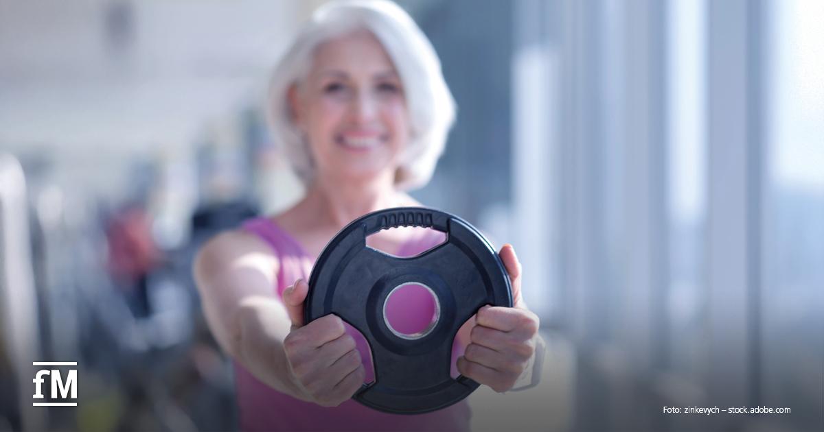 Ältere Frau im Fitnessstudio hält mit ausgestreckten Armen eine schwarze Gewichtsscheibe, Hintergrund mit Trainingsgeräten ist unscharf.