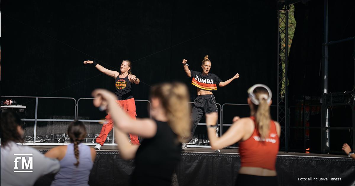 Zwei Trainerinnen führen eine Zumba-Choreografie auf einer Bühne an, während Teilnehmende vor der Bühne mittanzen.