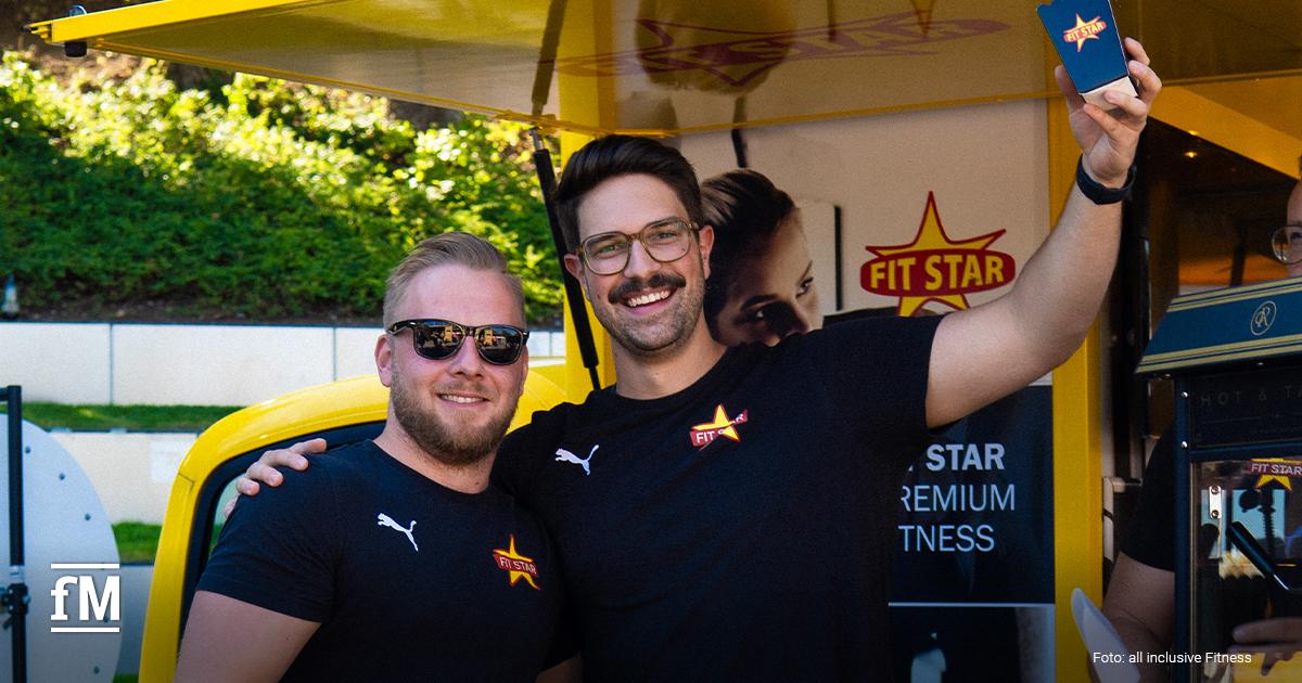 Zwei Männer in schwarzen FIT-STAR-Shirts posieren lachend vor einem gelben Stand.