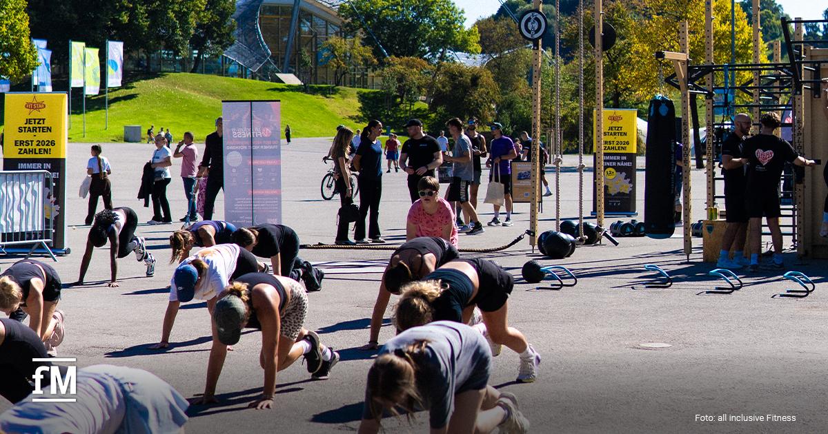 Mehrere Personen trainieren gemeinsam auf dem Boden, während andere im Hintergrund bei Outdoor-Fitnessübungen zusehen.