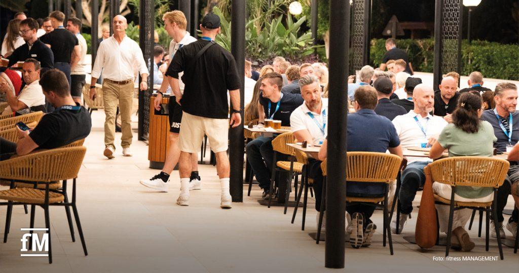 Menschen sitzen in einem offenen Restaurantbereich an Tischen und unterhalten sich, umgeben von Pflanzen.