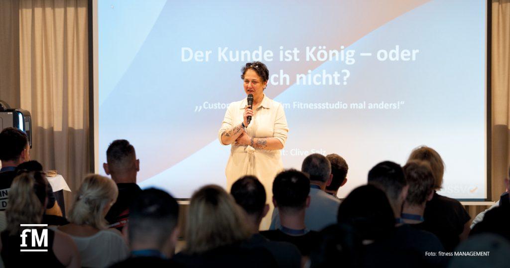 Eine Frau spricht mit Mikrofon vor Publikum, im Hintergrund eine Präsentationsfolie mit Vortragstitel.