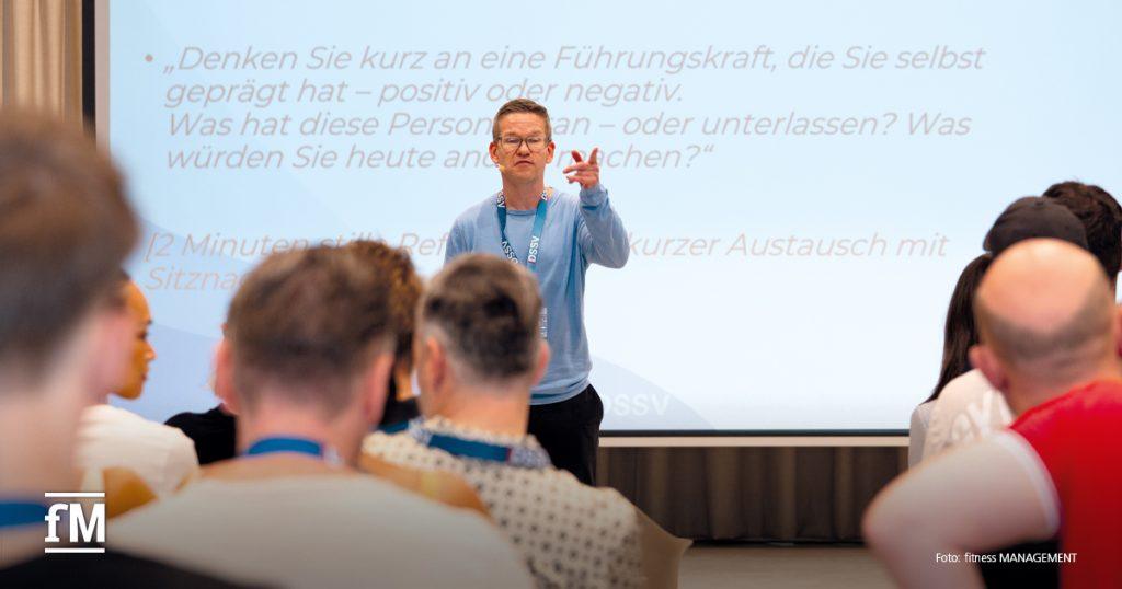 Referent spricht vor Publikum mit ausgestrecktem Arm, Hintergrund zeigt Vortragstext auf Leinwand.