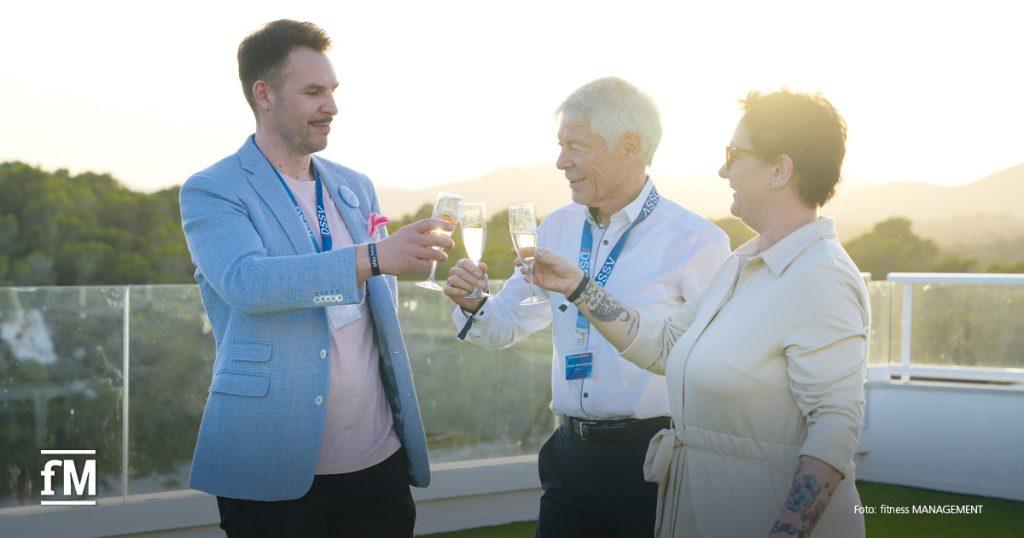 Drei Personen stoßen mit Sektgläsern auf einer Dachterrasse vor Landschaft im Abendlicht an.