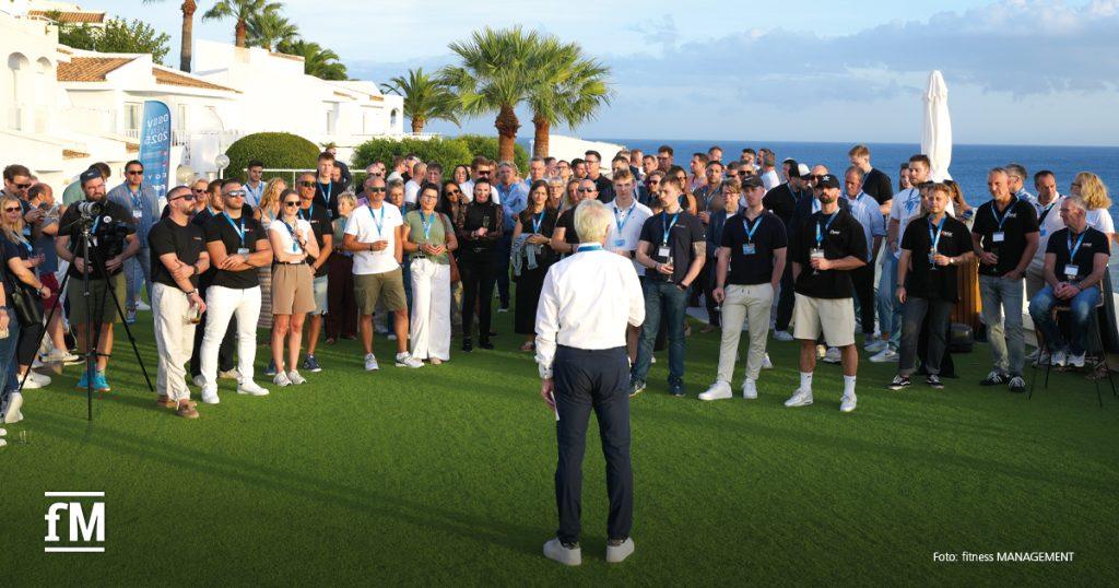 Person spricht vor einer großen Menschenmenge auf einer Wiese mit Meerblick im Hintergrund.