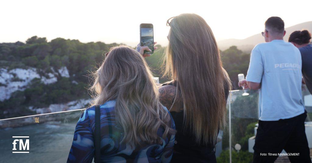Zwei Frauen fotografieren mit einem Smartphone den Sonnenuntergang über der Küste.