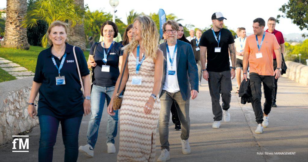 Gruppe von Personen geht auf einem gepflasterten Weg durch eine sonnige Hotelanlage mit Palmen.