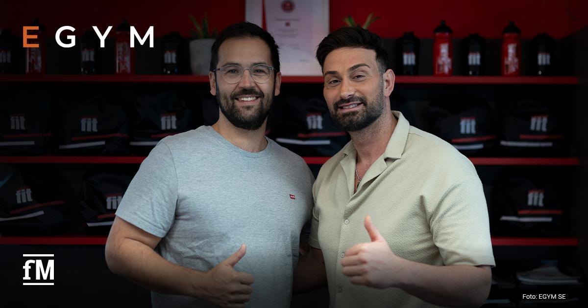 Zwei Männer stehen lächelnd am Service-Point eines Fitnessstudios und zeigen den Daumen nach oben, im Hintergrund Regale mit Trinkflaschen.