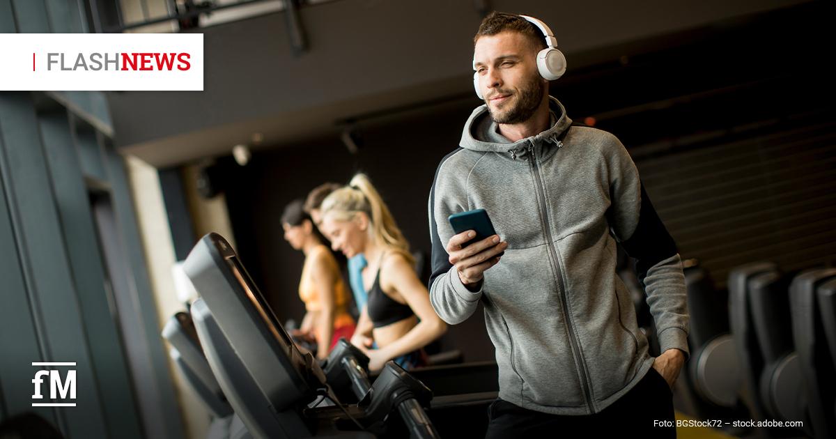 Fitness Flash News #01/2026 Mann mit grauem Hoodie und Kopfhörern läuft auf einem Laufband im Fitnessstudio und hält ein Smartphone in der Hand.