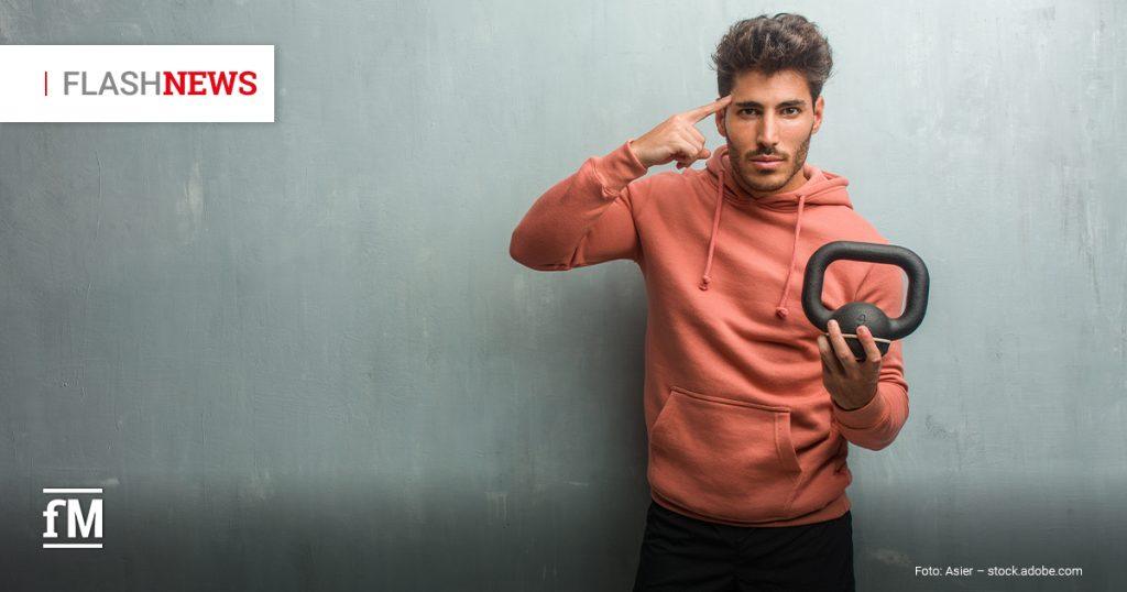 Ein junger Mann in Sportkleidung steht vor einer grauen Wand, trägt einen rostroten Hoodie und schwarze Shorts. In der rechten Hand hält er eine kleine Kettlebell, mit der linken Hand zeigt er nachdenklich auf seinen Kopf.