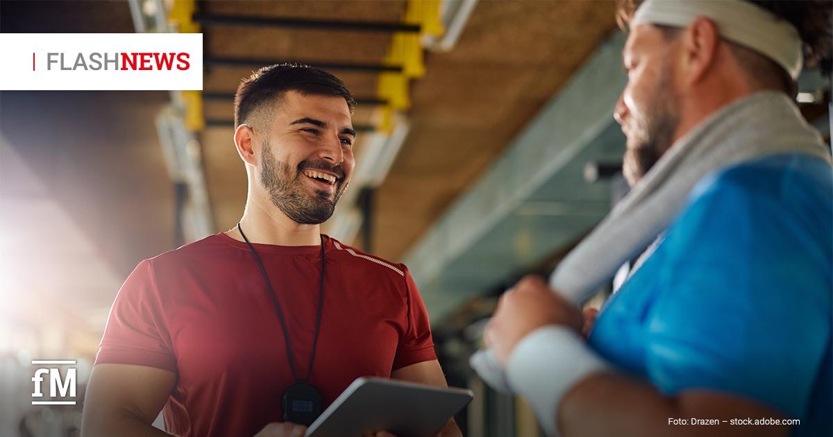 Ein junger Personal Trainer im roten Shirt hält ein Tablet und lacht, während er mit einem älteren Mann im blauen Shirt und Stirnband spricht; sie stehen in einem Fitnessstudio.