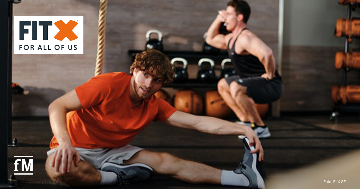 FitX Turnecke Zwei Trainierende in einem FitX Studio: vorne ein Mann im orangefarbenen Shirt beim Dehnen am Boden, hinten ein Mann bei einer Kniebeuge mit Gewichten.