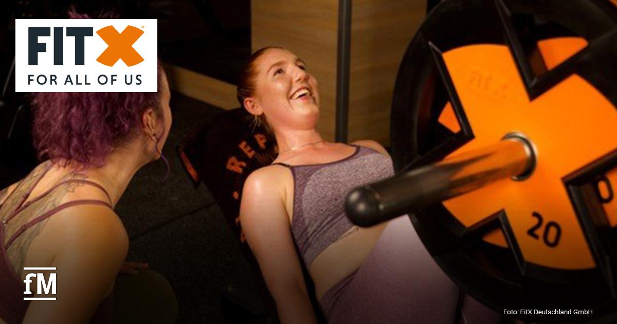 In einem FitX-Studio trainieren zwei Frauen an einer Beinpresse mit orangefarbenen Gewichtsscheiben. Eine liegt auf dem Gerät, die andere motiviert sie lächelnd von der Seite. Das FitX-Logo ist oben links auf weißem Hintergrund.