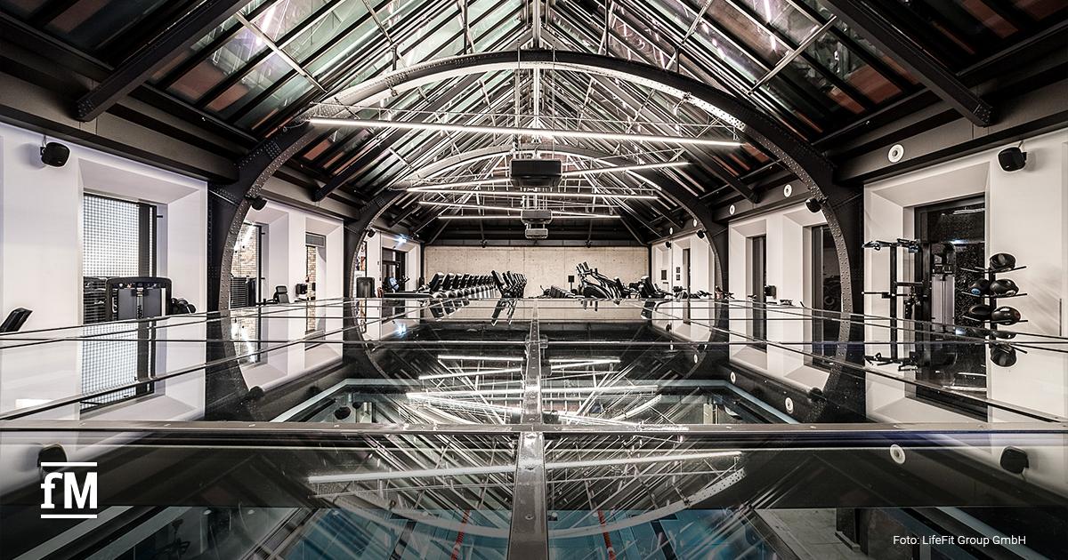 Großer lichtdurchfluteter Fitnessraum mit Glasdach, Glasboden, darunter ein Pool und Trainingsgeräten im Hintergrund.