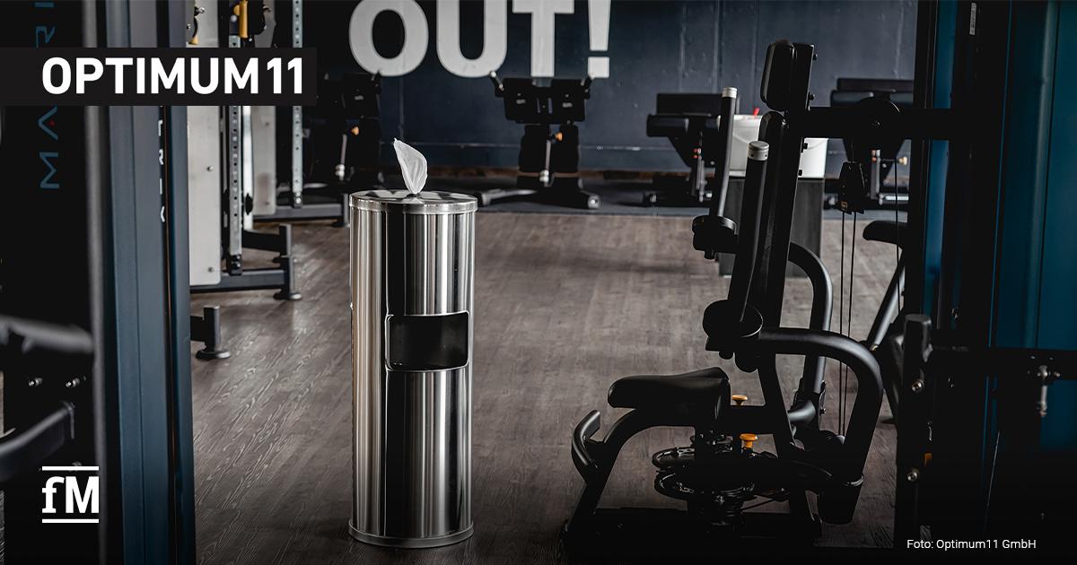 Edelstahlspender von Optimum11 für Reinigungstücher steht in einem Fitnessstudio zwischen Kraftgeräten auf einem Holzfußboden.