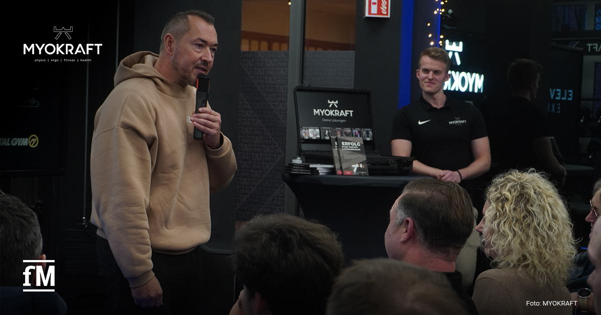 Ein Mann im Hoodie hält ein Mikrofon vor das Publikum. Rechts davon steht an einem Bartisch ein weiterer Mann. Auf dem Tisch steht ein aufgeklapptes Notebook mit dem MYOKRAFT-Logo und Flyern.